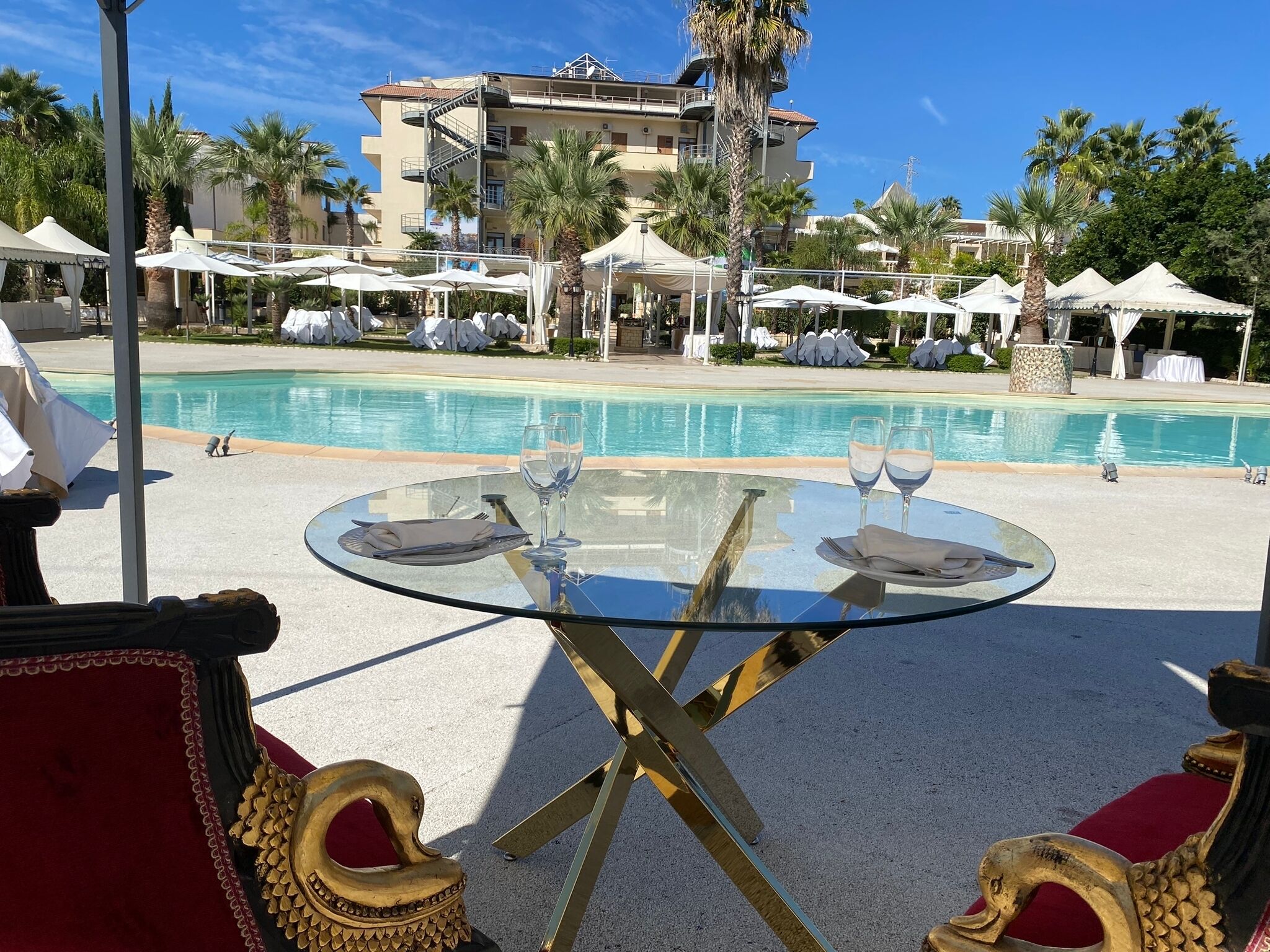 A poolside area with a glass table set for two, surrounded by red and gold ornate chairs.