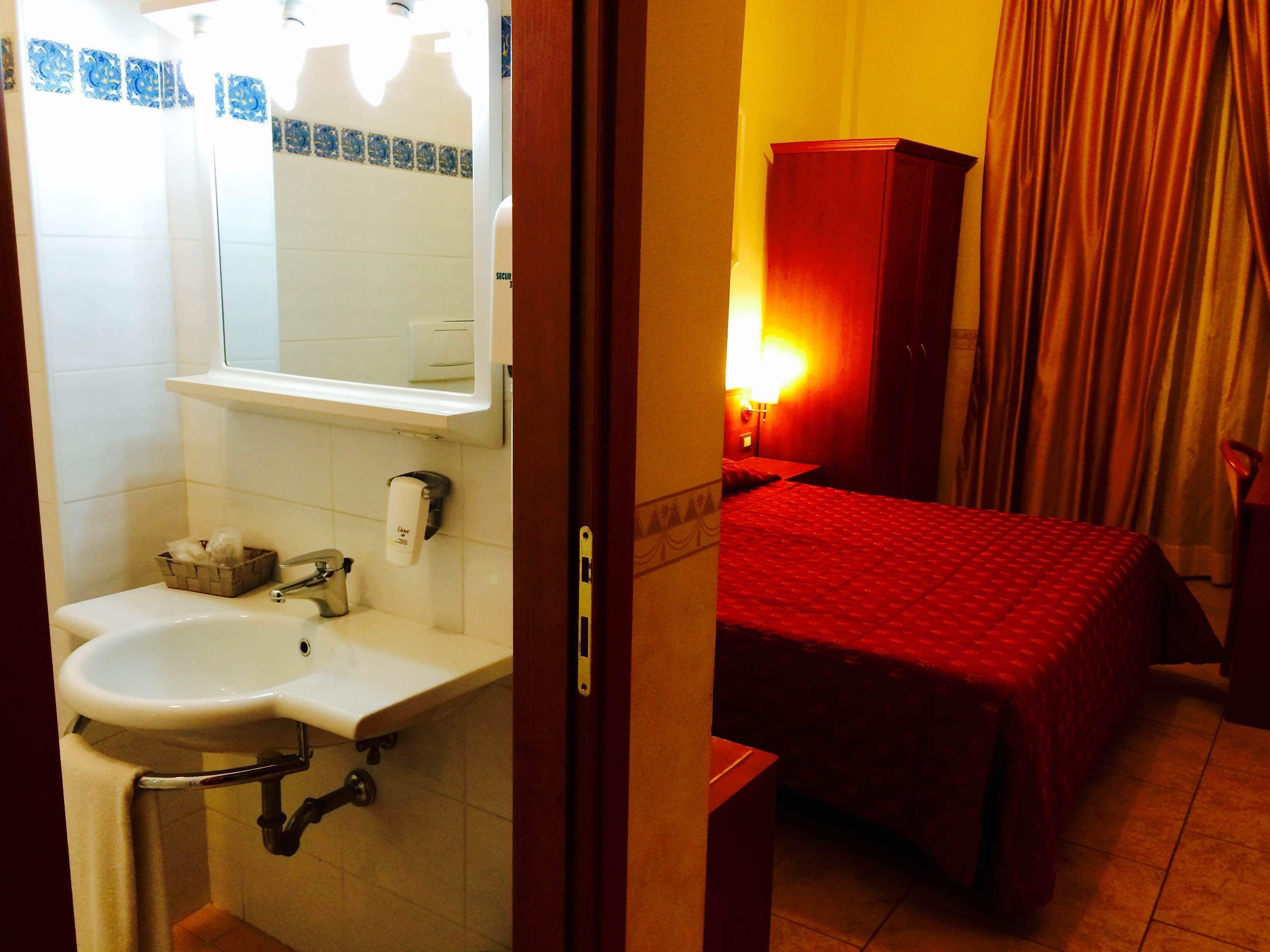 A hotel room with a bathroom featuring a sink, mirror, and towel rack. There is a bed with a red bedspread and a nightstand with a lamp.