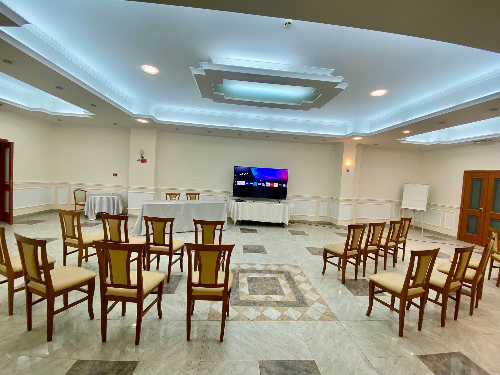 A conference room with a large flat-screen TV, multiple tables with white tablecloths, and rows of wooden chairs.