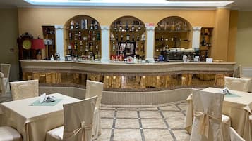 A bar area with a tiled floor, covered tables, and chairs, and a variety of bottles and glasses on shelves.