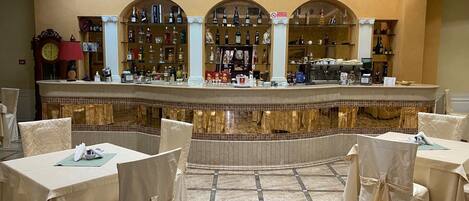 A bar area with a tiled floor, covered tables, and chairs, and a variety of bottles and glasses on shelves.