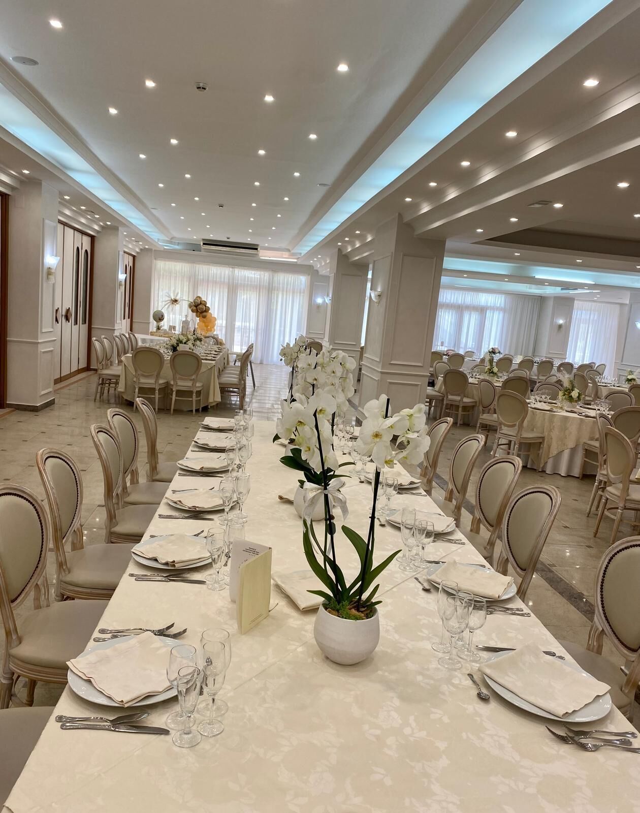 Een feestzaal met lange tafels, gedekt voor een formeel evenement, met witte bloemstukken en elegante tafeldecoraties.