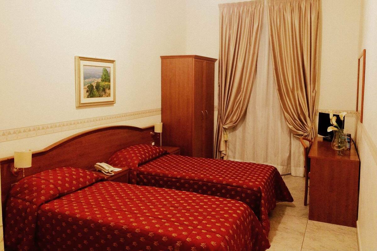 Habitación de hotel con dos camas, un armario de madera, un escritorio con lámpara y un cuadro en la pared.