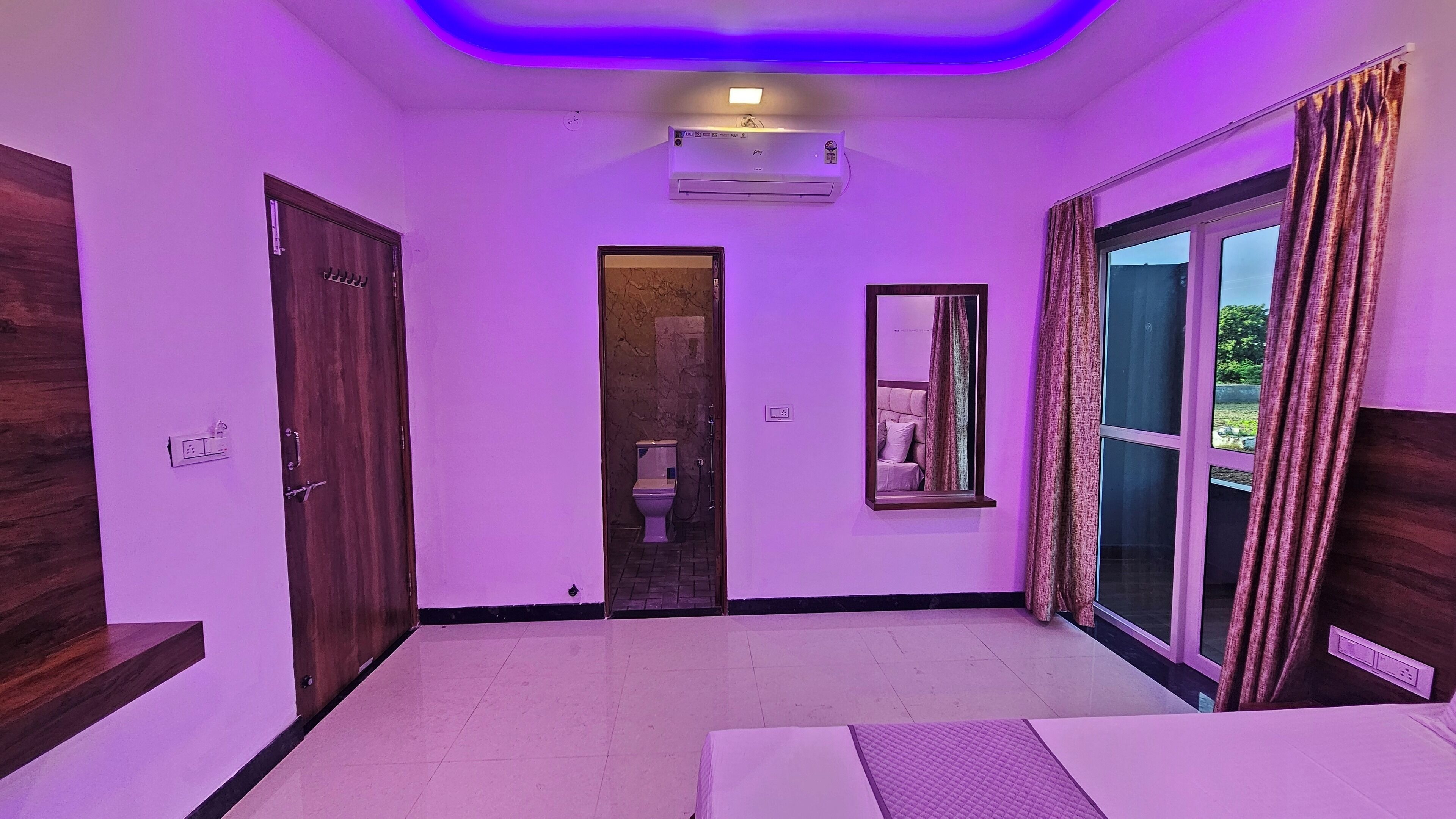 A hotel room with a large bed, a bathroom with a toilet and shower, a window with curtains, and an air conditioning unit.