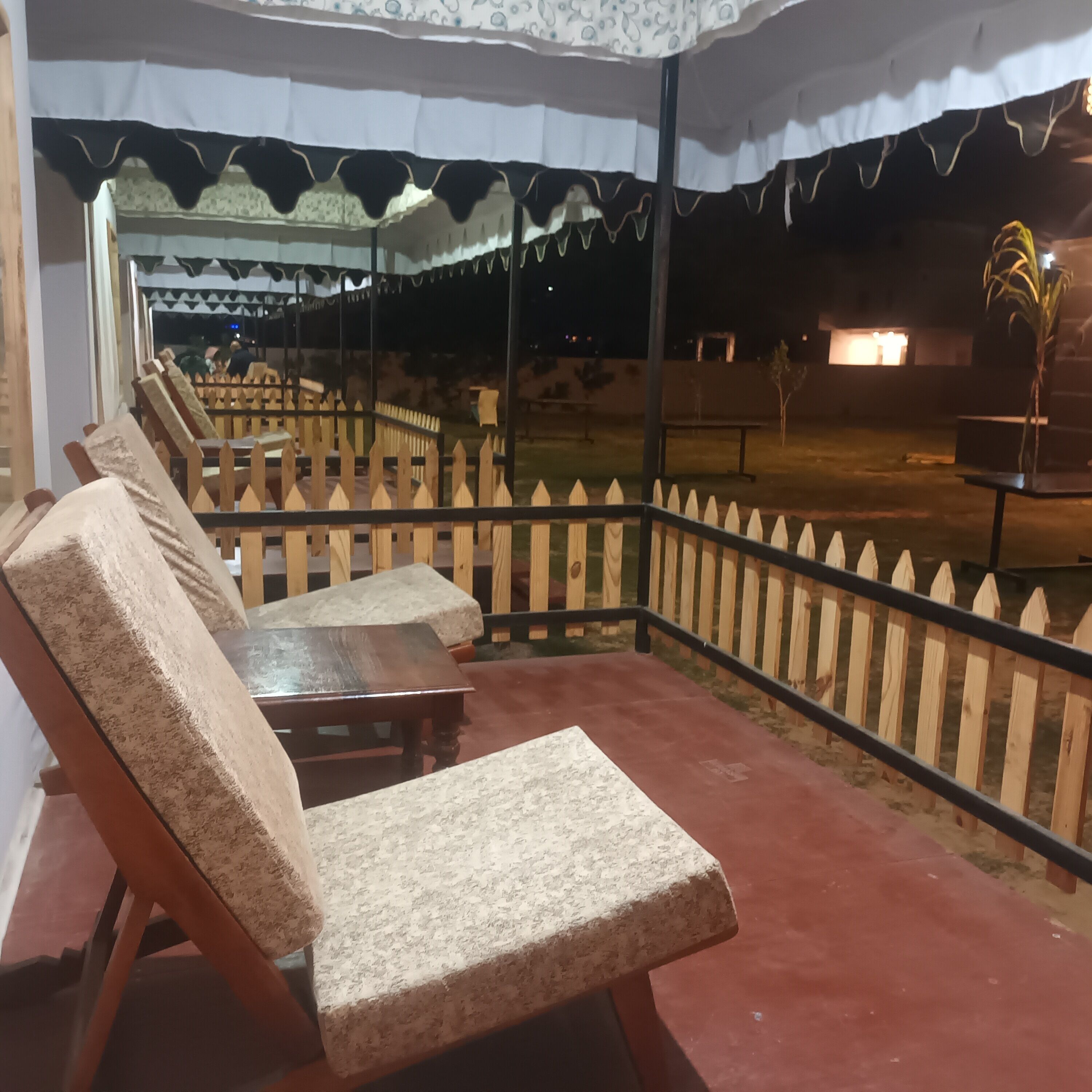 Un área de estar al aire libre cubierta con sillas y mesas de madera, una cerca y vistas al cielo nocturno.