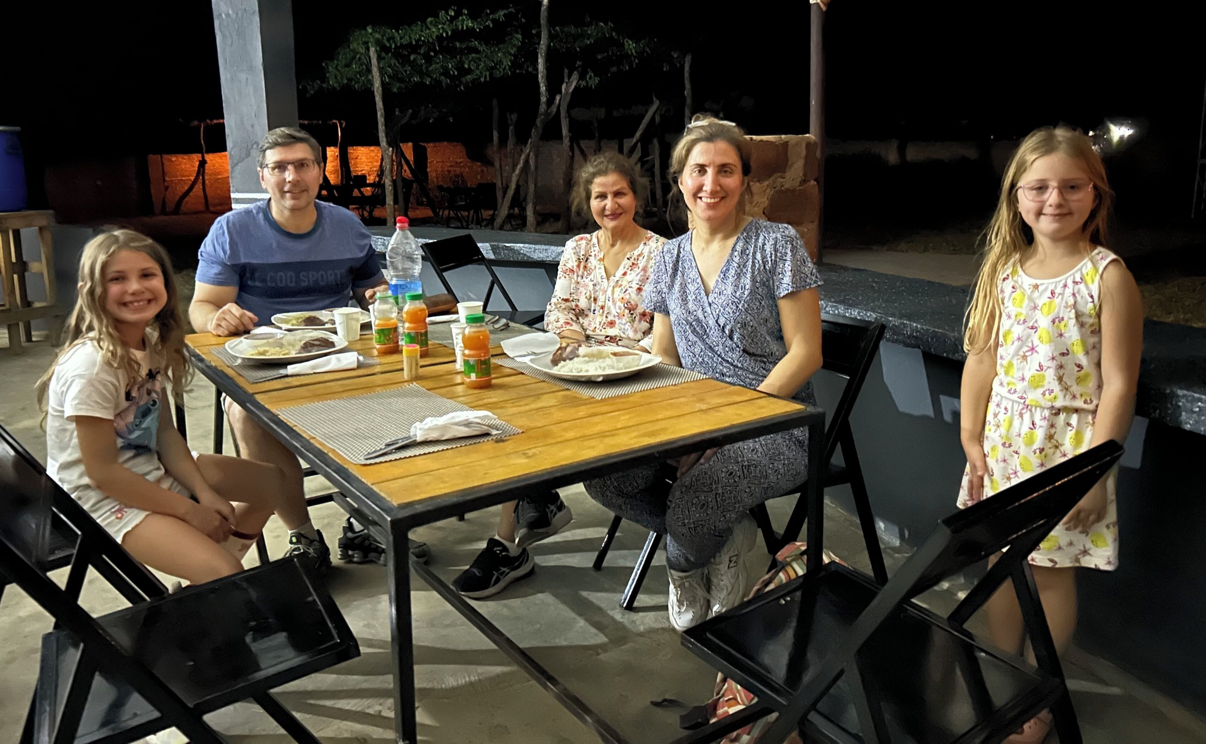 Una familia cenando al aire libre por la noche, con una mesa de madera, sillas y diferentes platillos.