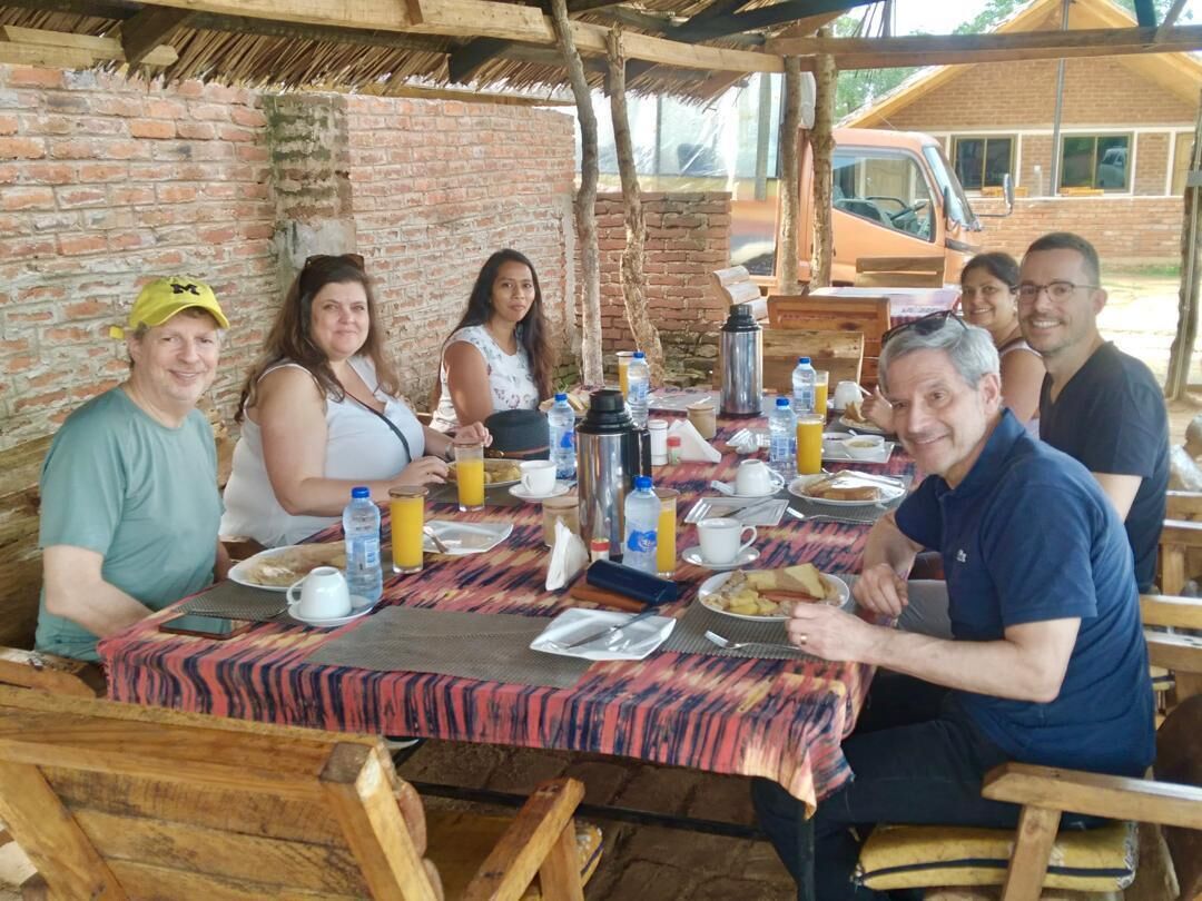Un grup de persones dinant a una taula a l'aire lliure, amb un mur de maó i un teulat de palla al fons.