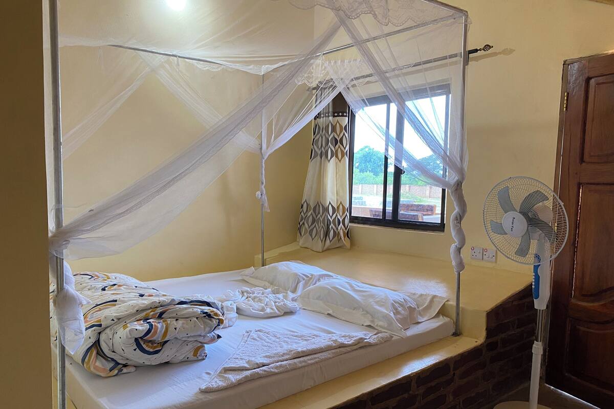 Un letto singolo con zanzariera, una ventola a lato del letto e una finestra con tende.