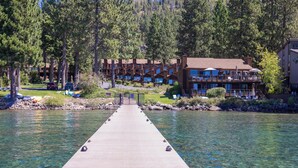 A wooden pier leading to a lodge with multiple balconies and rooms, surrounded by trees and a clear lake.