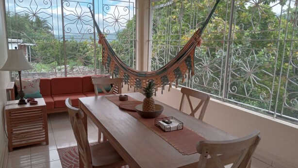 A dining area with a table, chairs, a hammock, and a view through large windows. - Enjoy Beverly Hills, Kingston, Jamaica, 4 bdrms, AC, WiFi, Spacious, Quiet, Safe (Kingston)