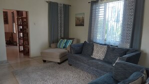 A living room with a sofa, ottoman, and a rug. There is a door leading to another room and a window with curtains. - Enjoy Beverly Hills, Kingston, Jamaica, 4 bdrms, AC, WiFi, Spacious, Quiet, Safe (Kingston)