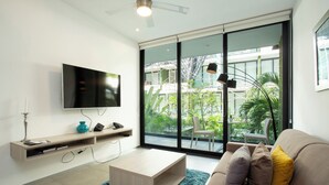 A modern living room with a flat-screen TV, a wooden coffee table, a beige sofa with cushions, and a large sliding glass door leading to a balcony with plants and a view of a building. - Lovely One Bedroom With Amazing Terrace View!! (Playa del Carmen)