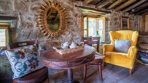 A rustic room with a stone wall, wooden ceiling beams, a round wooden table with a tray of white teacups, a yellow armchair with a pillow, and a mirror with a sunburst frame. - Cosy cottage in Los Valles Pasiegos, Cantabria: Lo Lucas 1920 (San Roque de Riomiera)