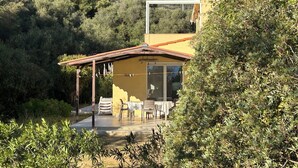 Una casa de dos pisos con un patio, rodeada de abundante vegetación y colinas al fondo.
