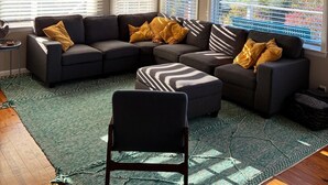 A modern living room with a sectional sofa, a chair, and a ceiling fan. Large windows provide natural light and a view of trees.