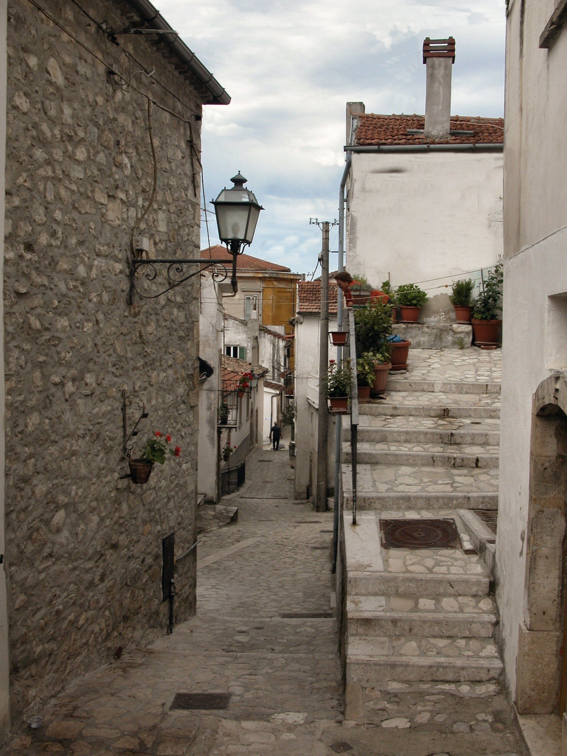 A narrow cobblestone street flanked by historic stone buildings with terracotta roofs, a street lamp, and potted plants.