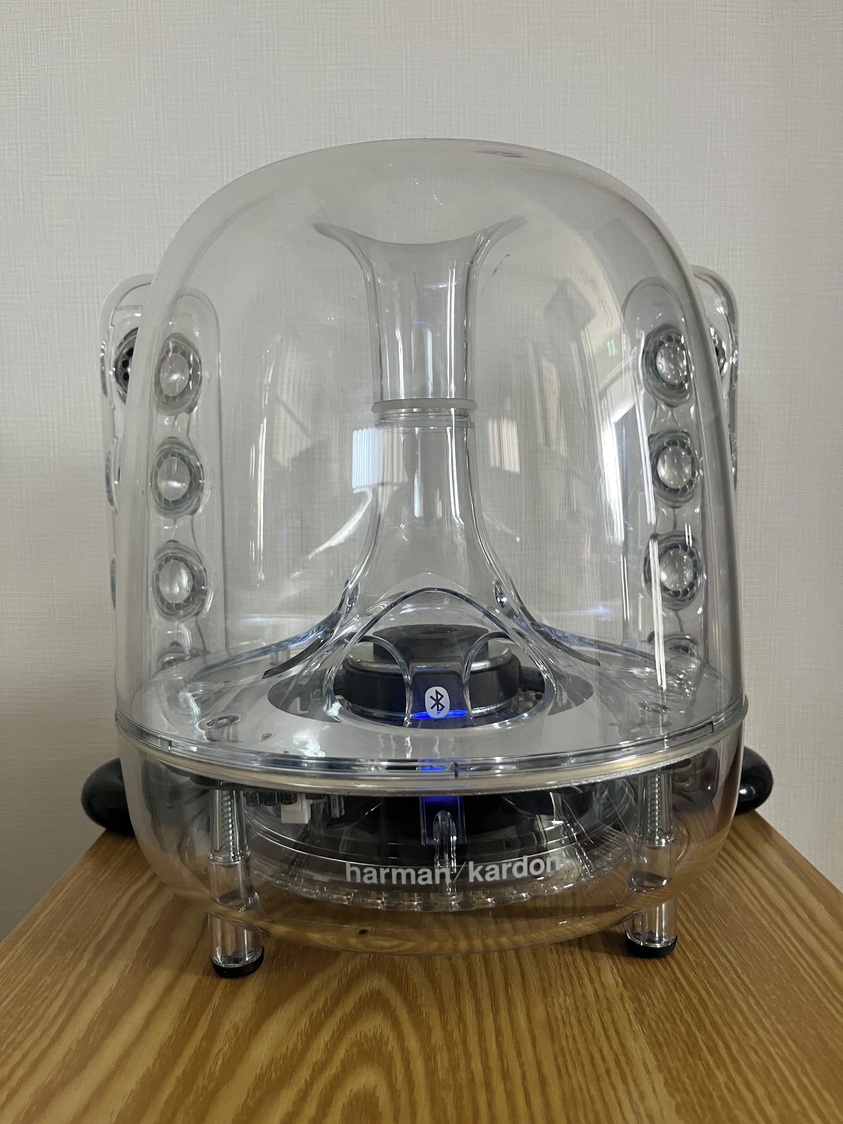 Un oggetto a forma di cupola trasparente con diverse aperture circolari, con la scritta "harman kardon", su una superficie in legno.