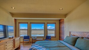 Ein Schlafzimmer mit einem großen Bett, einem Fernseher, einem Schreibtisch und zwei Stühlen. Drei Fenster bieten Blick auf eine gebirgige Landschaft.
