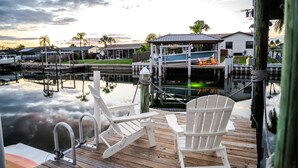 A wooden dock with two white Adirondack chairs, a metal railing, and a view of a marina with boats and houses in the background. - Waterfront | Dock, Htd Pool, Hot Tub & Game Room (Apollo Beach)