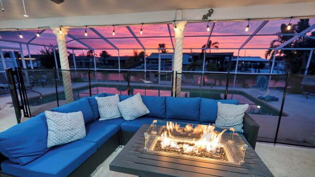 A covered outdoor seating area with a fire feature and a view of the pool and sunset. - Waterfront | Dock, Htd Pool, Hot Tub & Game Room (Apollo Beach)