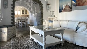 Uma sala de estar espaçosa com um sofá branco, uma mesa de centro de vidro e um candelabro. Há uma escada que leva a outro cômodo, e um quadro de um navio na parede.