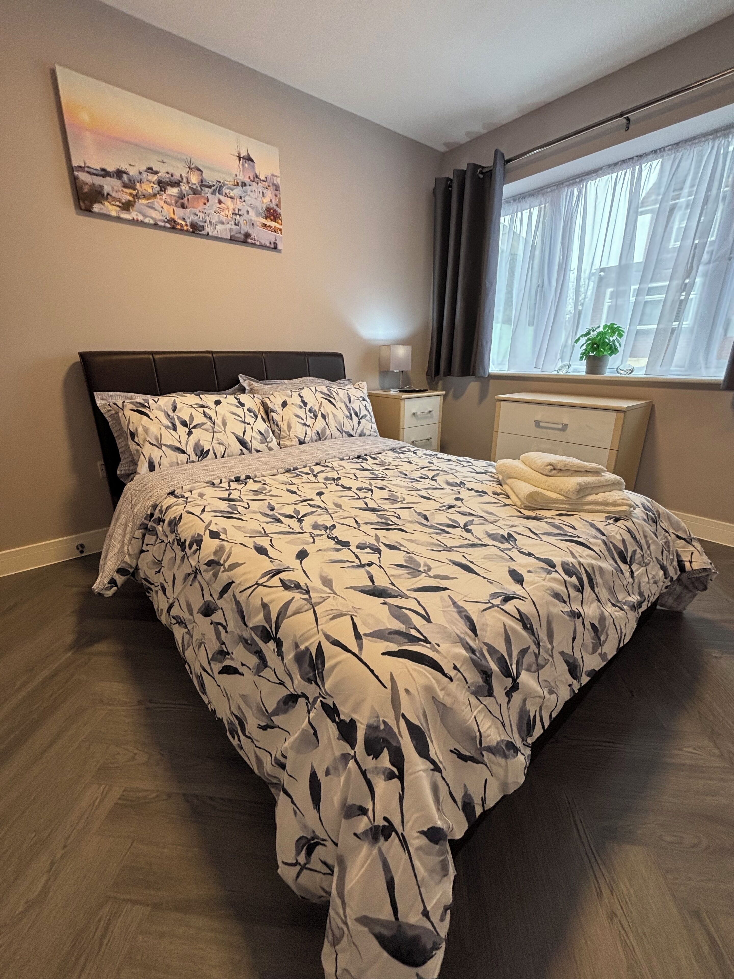 A bedroom with a large bed, a nightstand, a dresser, a window with curtains, and a painting on the wall.