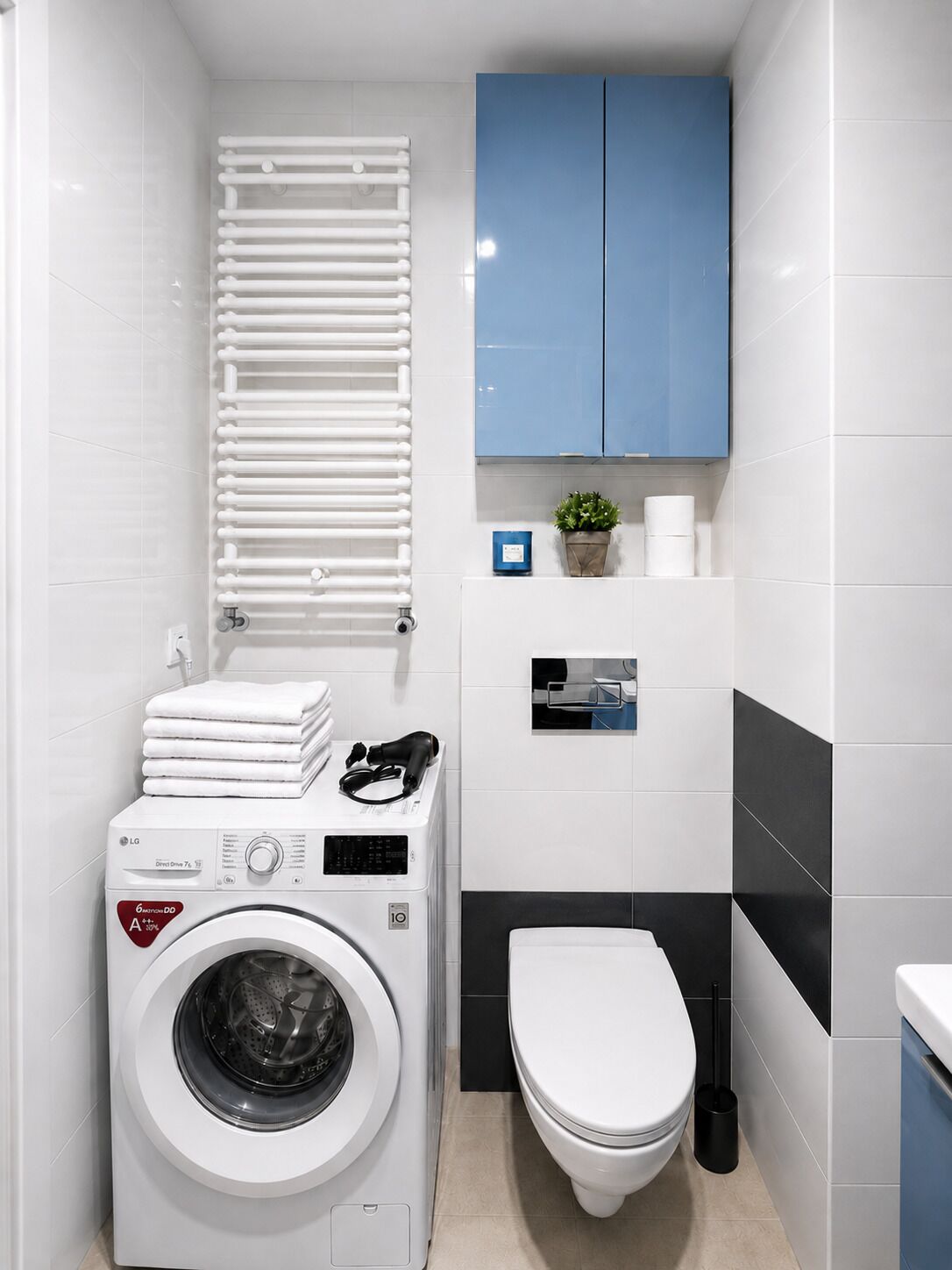 A compact laundry room with a front-loading washing machine, a toilet, a towel rack, and a small shelf with a plant and a photo.