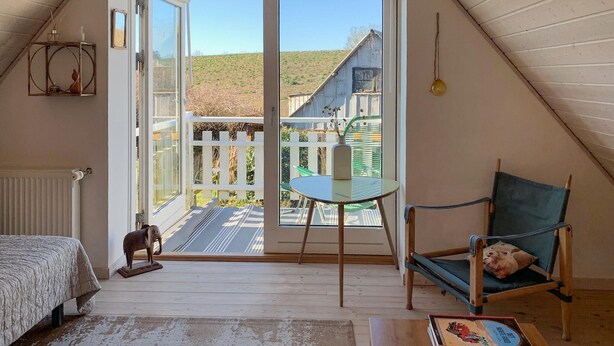 Ein gemütliches Dachzimmer mit einem Bett, einem kleinen Tisch, einem Stuhl und Blick auf eine Holzterrasse und ein Haus durch ein großes Fenster.