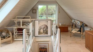 Ein gemütliches Dachzimmer mit schrägen Wänden, einer Holzleiter, einem gerahmten Bild, einem Bücherregal und einem Korbstuhl.