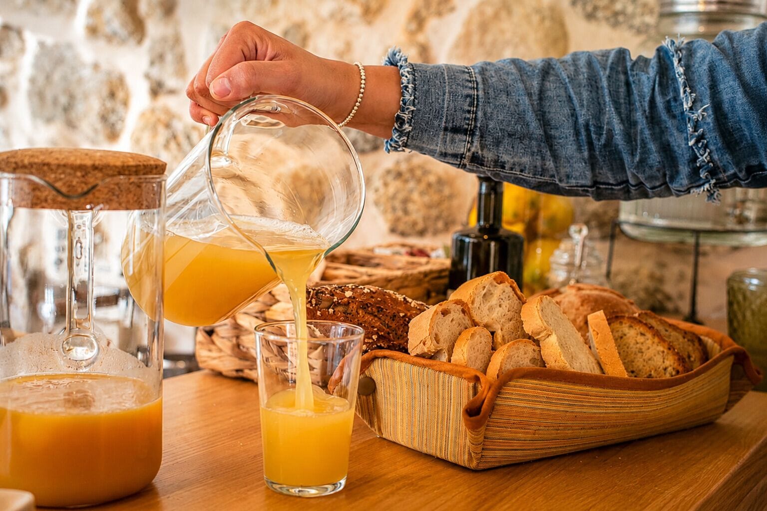 Una persona sirve jugo de naranja en un vaso, con pan y una cesta sobre una mesa de madera.