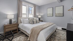 A bedroom with a bed, nightstands, a chair, a window with curtains, and two wall-mounted artworks.