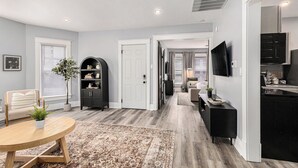A modern living room with a round wooden table, a sofa, and a large rug. There is a black entertainment unit and a white door leading to another room. - Remodeled, Washer, Dryer, Mins to Beach & Downtown (Michigan City)