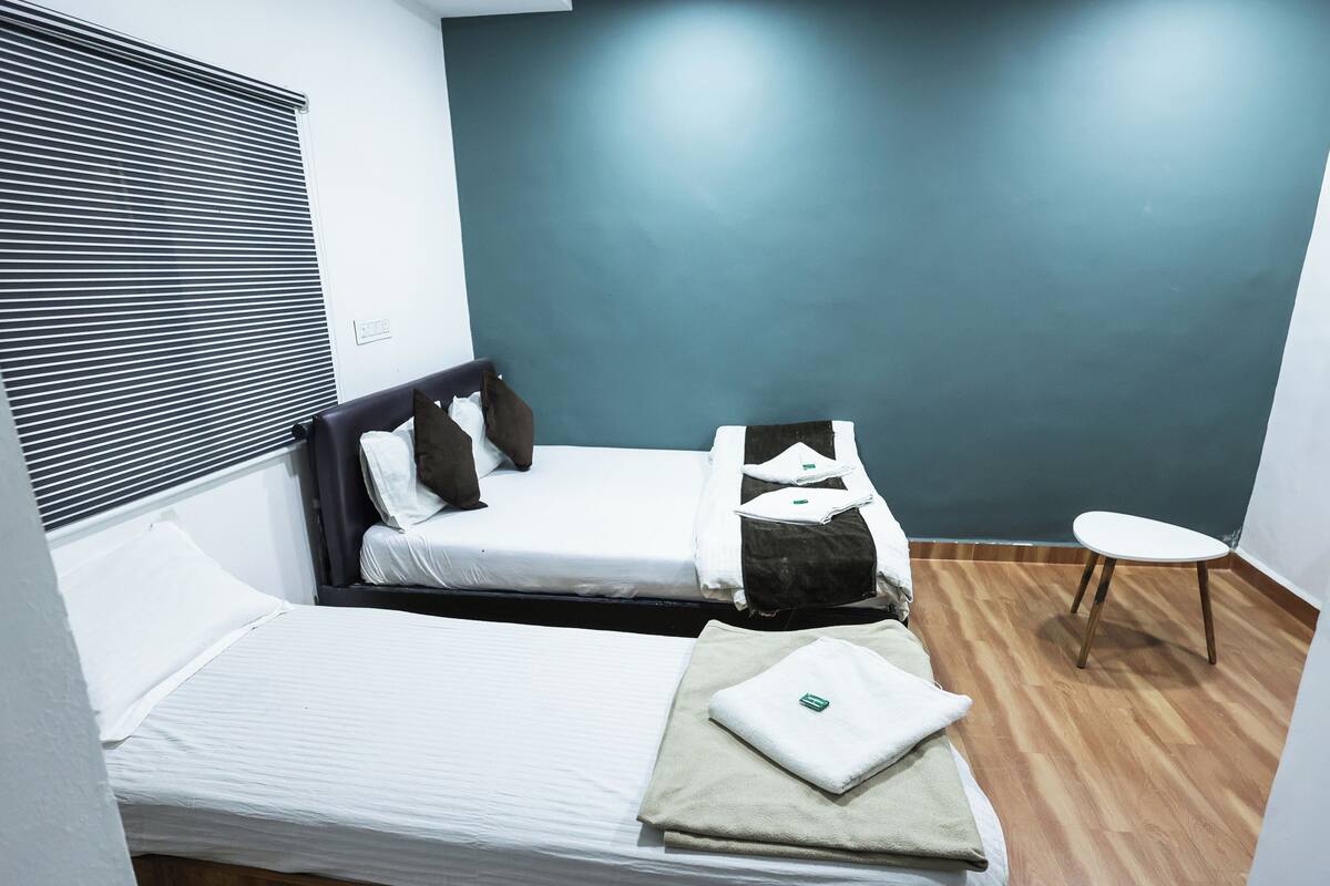 Une petite chambre propre, avec deux lits, une petite table et une fenêtre avec des stores.