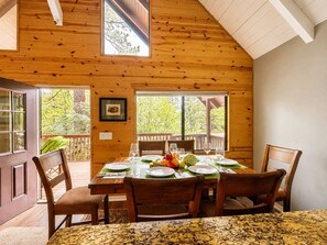 A dining area with a wooden table set for four, surrounded by chairs, with a view of trees through large windows.