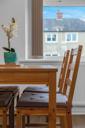 A wooden dining table with cushioned chairs, a teal potted orchid, and a view of a building through a window with a curtain. - Vacant Nests | The Grampian | Free Parking (North Lanarkshire)