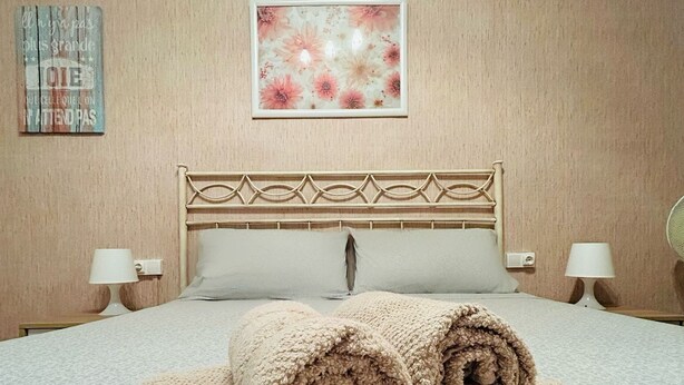 A neatly made bed with rolled towels, a decorative headboard, and a floral artwork above it.