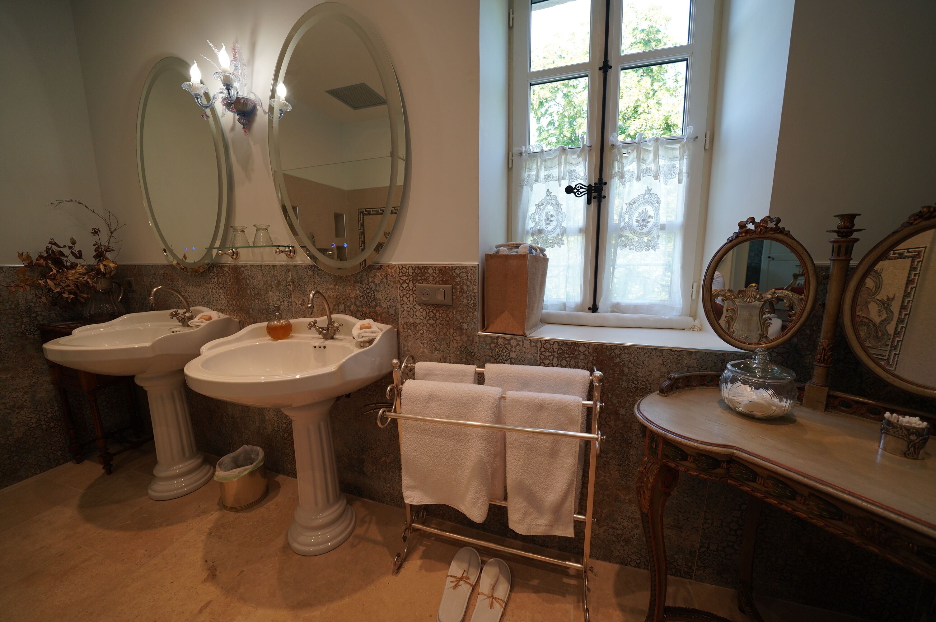 A bathroom with two pedestal sinks, a vanity with a mirror, a towel rack, and a window with curtains.