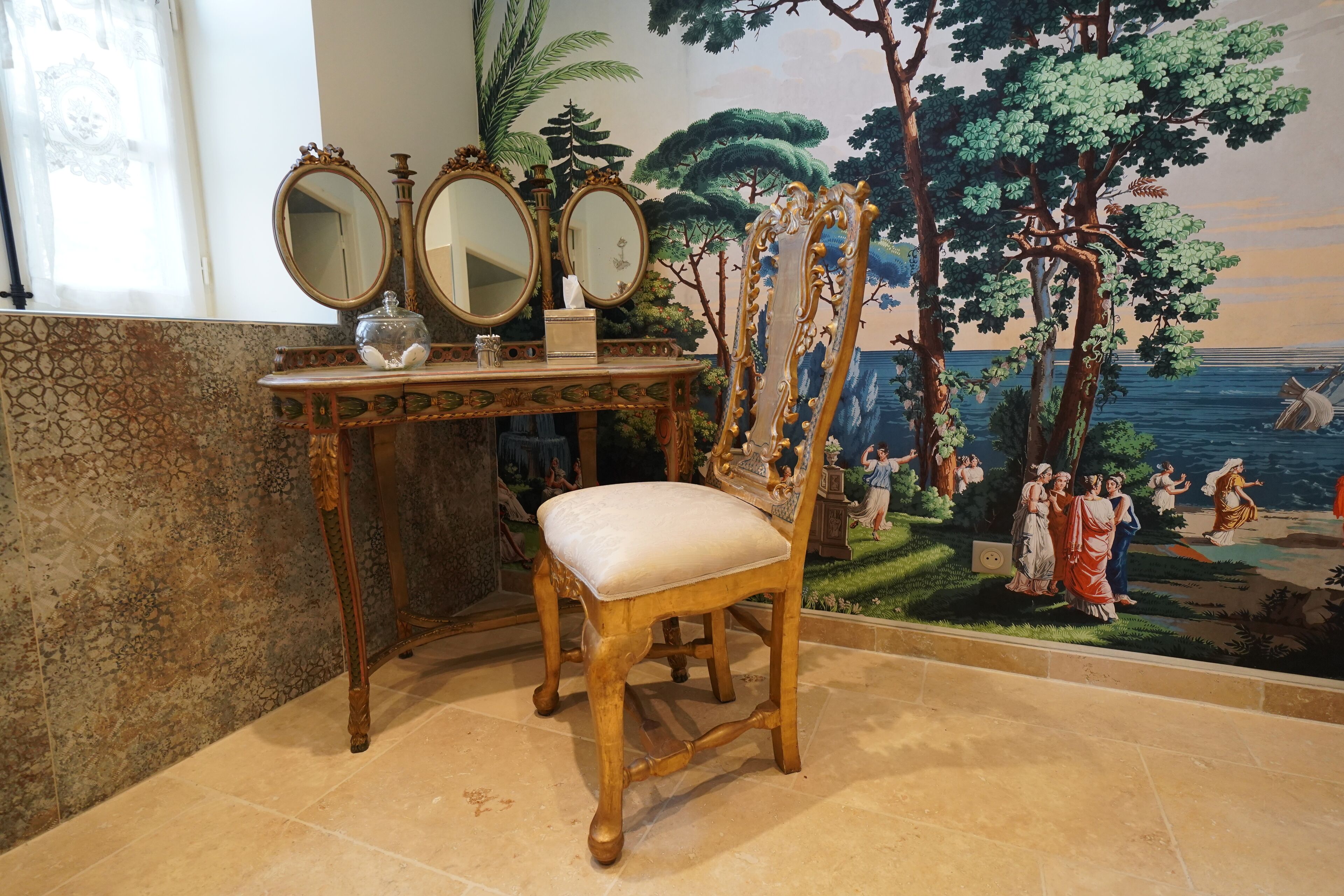 Une pièce comprenant une chaise en bois, une console surmontée de trois miroirs et une fresque murale représentant une scène de plage.