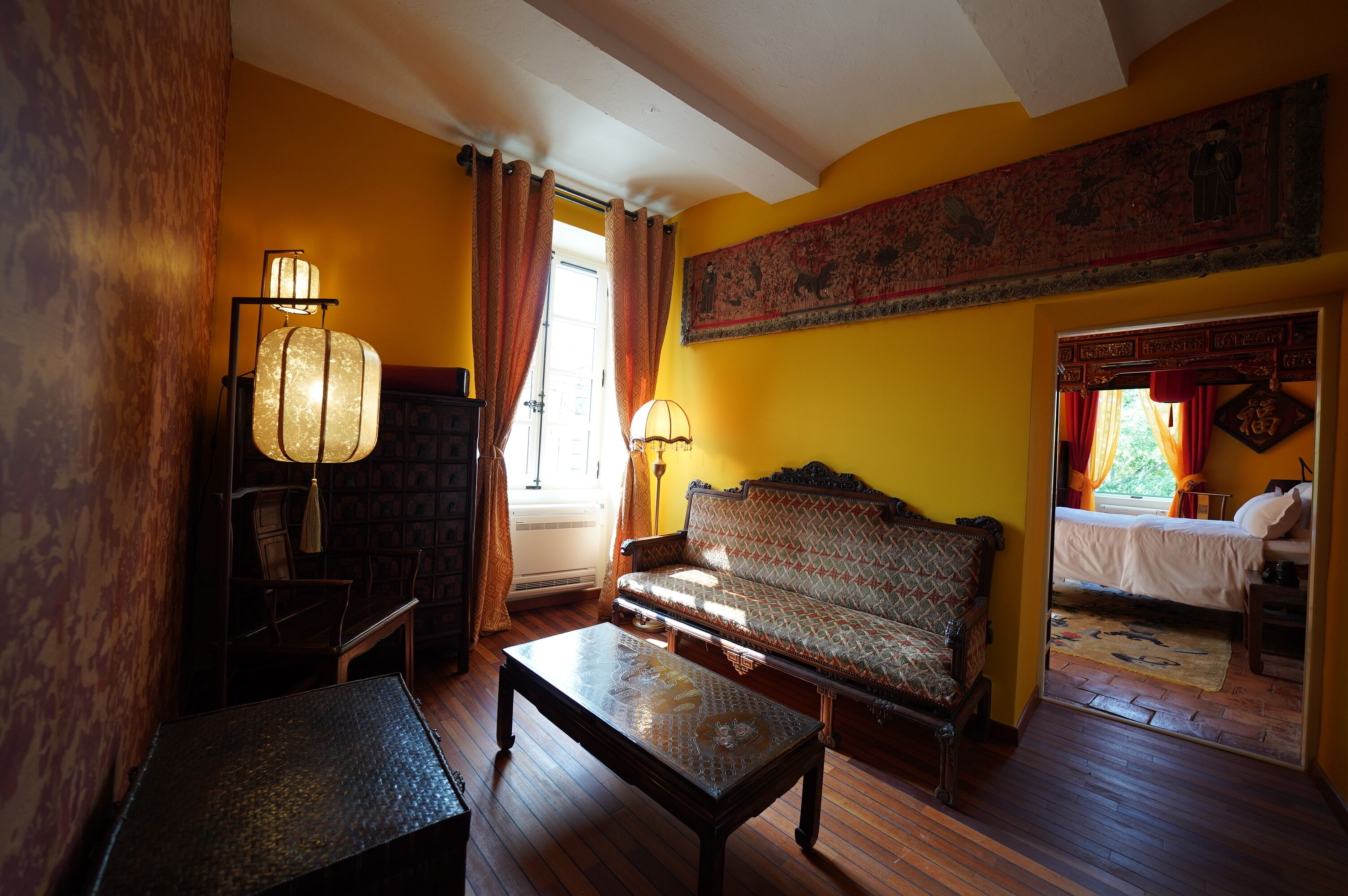 A room with a wooden floor, a sofa, a table, and a lamp. There is a window with curtains and a painting on the wall.