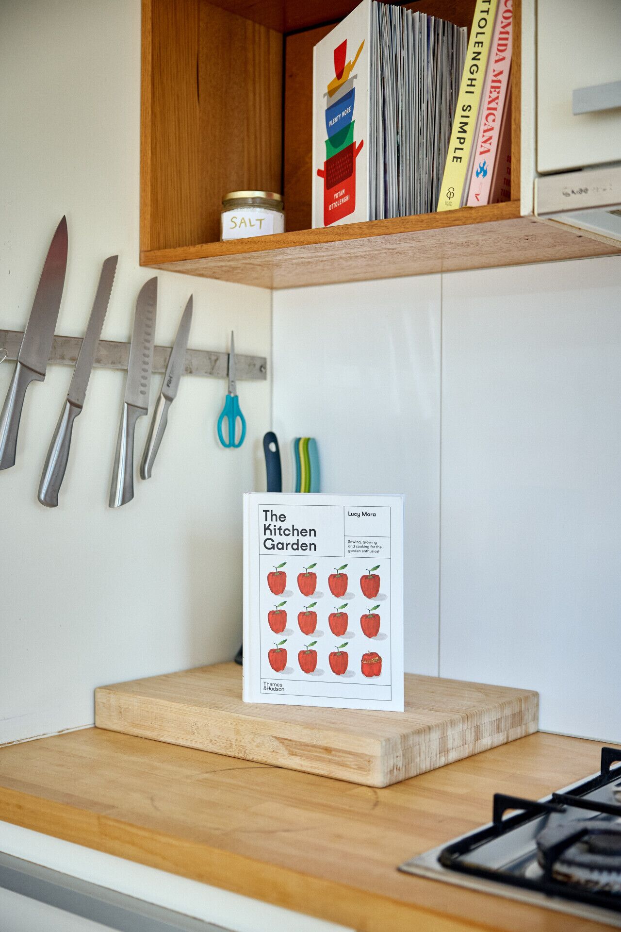 A kitchen counter with a book titled "The Kitchen Garden" by Lucy Mora, a salt container, and a set of knives.