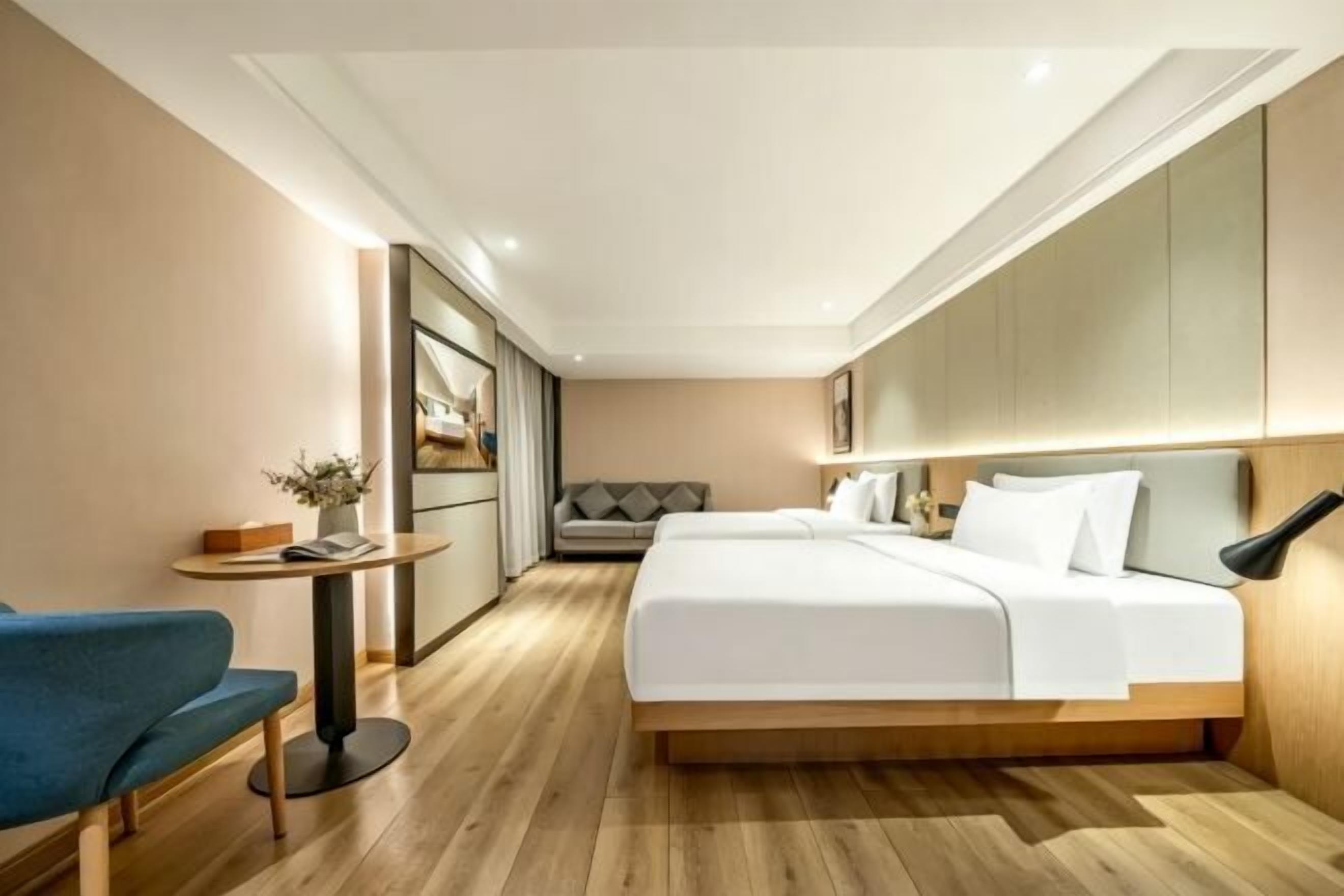 A modern hotel room with a bed, bedside table, chair, and a small table with a plant.
