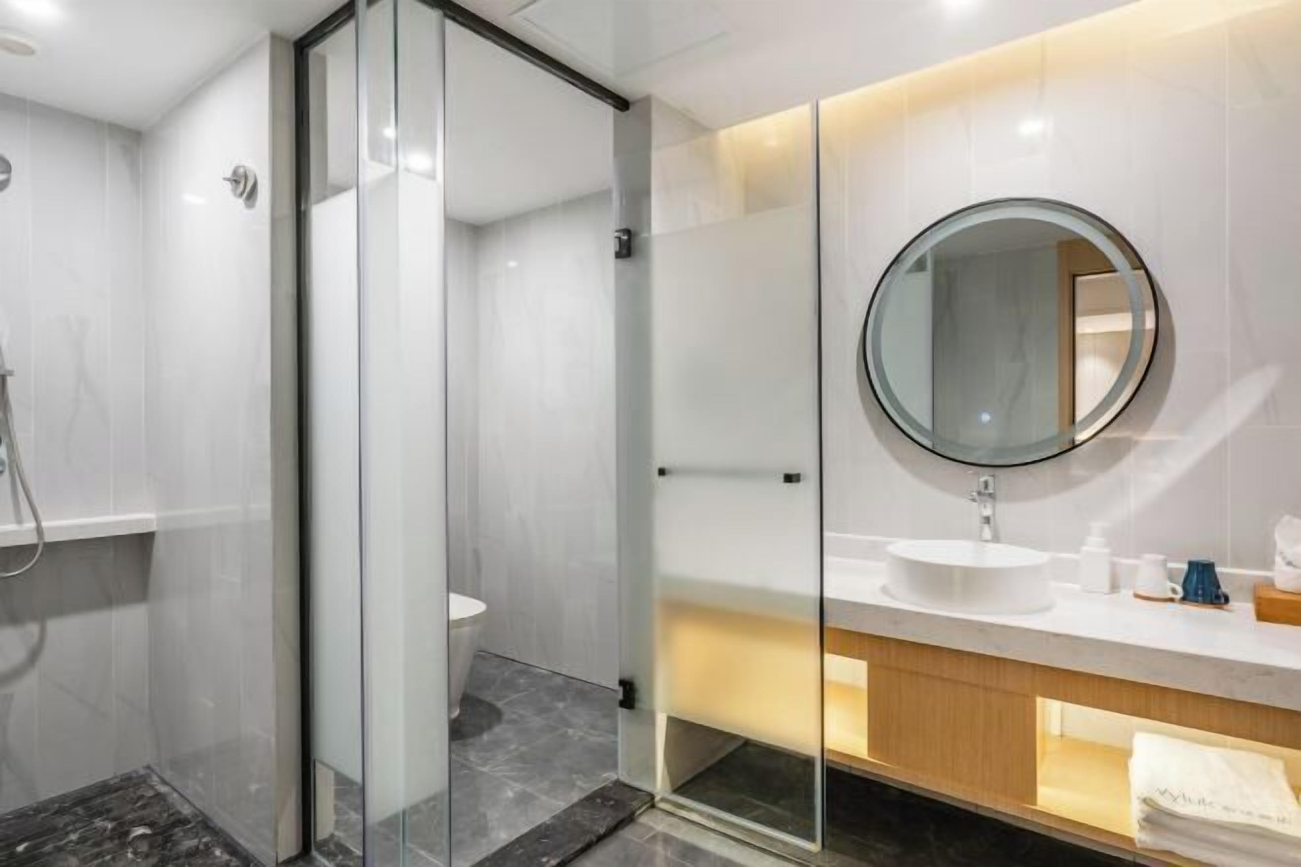 A modern bathroom with a glass shower enclosure, a round mirror, and a white sink with a wooden vanity.