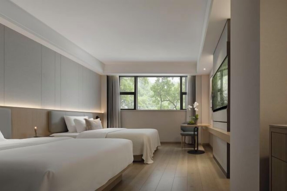 A hotel room with two beds, a desk, and a window with a view of trees.