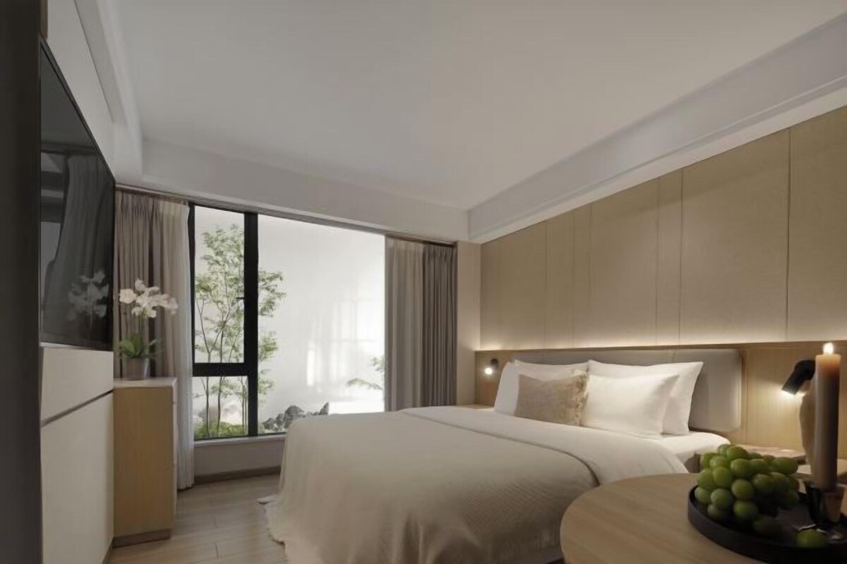 A modern hotel room with a large bed, a bedside table with a lamp, a window with curtains, and a view of the outdoors.