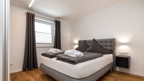A modern bedroom with a large bed, grey bedding, a window with curtains, a bedside table with a lamp, and a wooden floor.