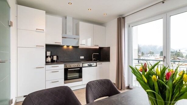 A modern kitchen with white cabinets, a dark countertop, and a dining area with two chairs and a table.