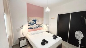 A hotel room with a bed, bedside lamps, a wall-mounted artwork, a bedside table, a fan, and a wardrobe.