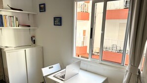 A room with a white desk, a laptop, a bookshelf, and a window with a view of a balcony.