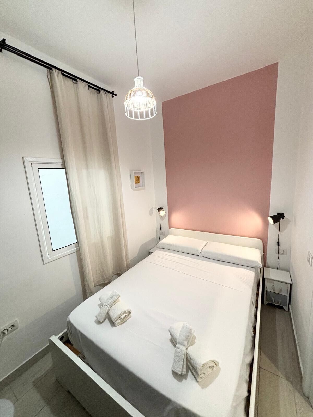 A neatly made bed with white linens and towels, a bedside table, a hanging light fixture, and a window with curtains.