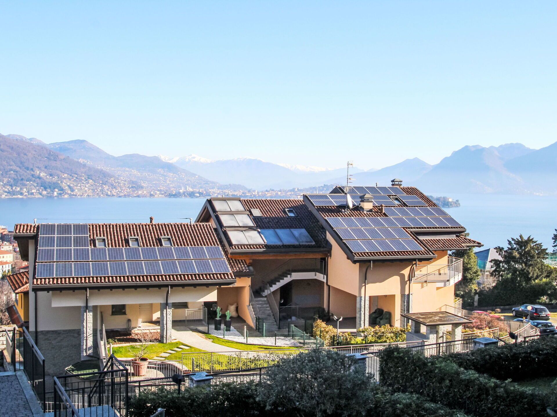 A residential building with solar panels on the roof, a garden, and a view of a lake and mountains.